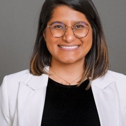 Photo of a South Asian woman with a cream jacket and a black top. She has shoulder length black and brown hair and is wearing round glasses.