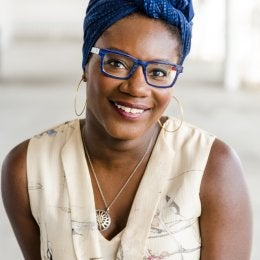 An image of a Black woman wearing a blue square-framed glasses, a blue headwrap, large gold hooped earrings, a nautilus necklace, and sleeveless dress. 