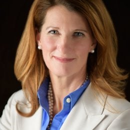 woman with brownish red hair wearing a blue shirt and white jacket.