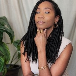 Amber is wearing a beige dress and seated in front of a beige background. Her locs are hanging and her chin is resting on her hand. 