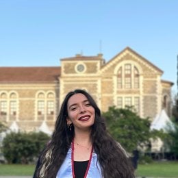 A woman smiling outdoors, wearing a graduation gown and stole, standing in front of a historic university building on a sunny day.