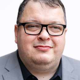 Everett wearing a grey suitcoat, with short brown hair and black thick rimmed glasses.