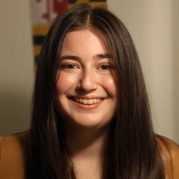 This is an image of Sivan Lurie. She is smiling and has a Maryland flag behind her