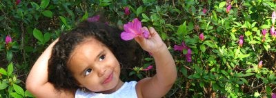 Young girl standing next to bushes looking at flower