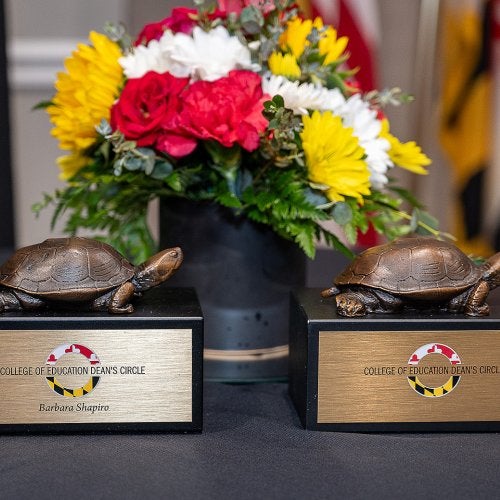 Two Dean's Circle trophies (with a bronze Testudo sitting on a black box) in front of a bouquet of yellow, red and white flowers
