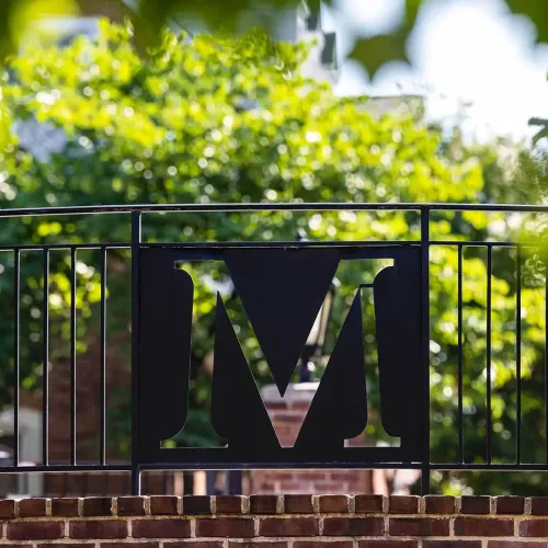 The letter M in a wrought-iron fence on the South Campus Commons, surrounded by brickwork and greenery.
