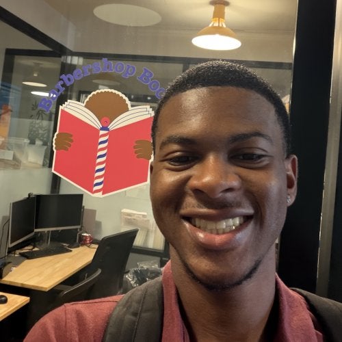 Emmanuel Dean '28 stands in front of an office with the "Barbershop Books" logo on the window, featuring a Black child reading a red book with a barbershop pole for a spine.