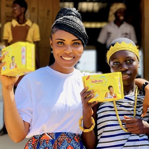 Joyce Arthur M.A. ’26, a Ph.D. student, stands with her arm around a girl in Ghana. They hold up yellow packages containing free menstrual products that Arthur distributes through her nonprofit, Araba Foundation International.
