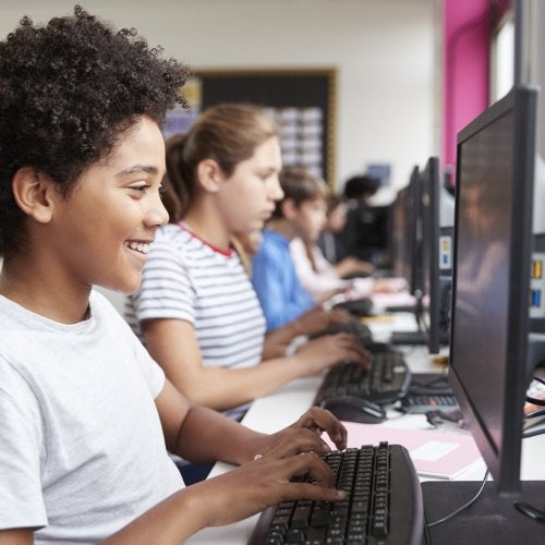 A middle school student smiles while working on a computer, in front of a line of other students at work on computers as well.