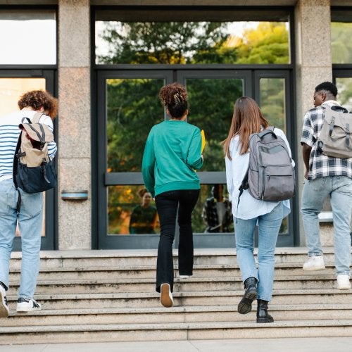 Four high school students wearing backpacks walk up steps into a school building.