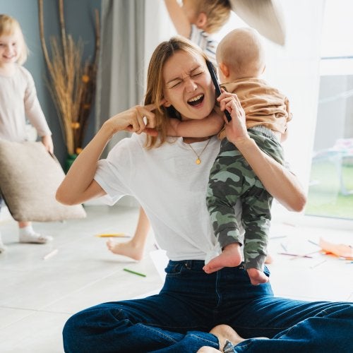 A mother puts her finger in her ear while holding a baby and a cell phone, as two children have a pillow fight in the background.