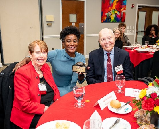 Patricia S. “Pat” Koskinen Ph.D. `75 and John A. Koskinen at a table with Dean Kimberly Griffin, who holds up a Dean's Circle trophy