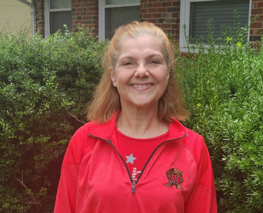 A woman wearing a red UMD logo jacket standing by trees smiling for a portrait. 
