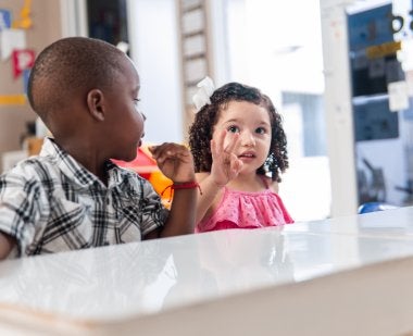 A little girl in a classroom holds up three fingers.