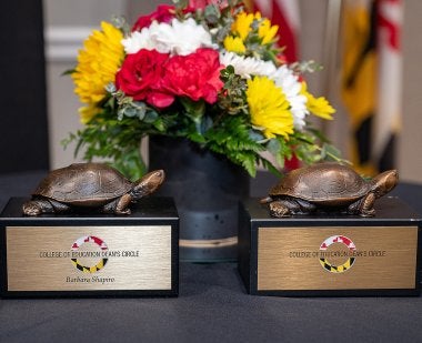 Two Dean's Circle trophies (with a bronze Testudo sitting on a black box) in front of a bouquet of yellow, red and white flowers