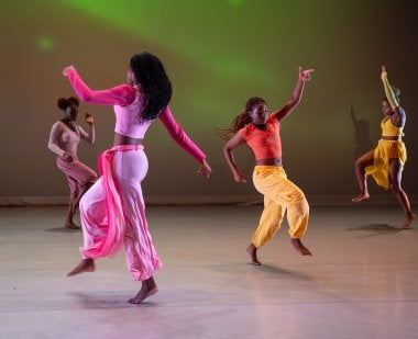 Four dancers dressed in bright colors dance across a stage.