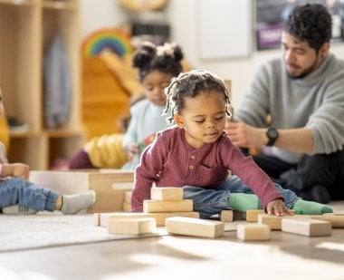 A preschool-aged boy plays with blocks, with two other children and a man in the background.