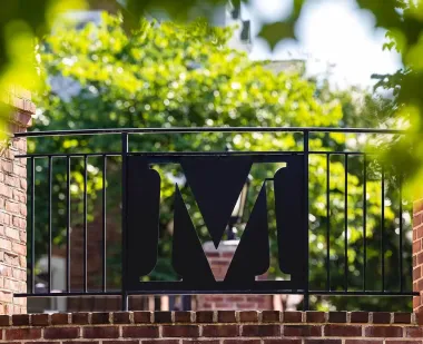 The letter M in a wrought-iron fence on the South Campus Commons, surrounded by brickwork and greenery.