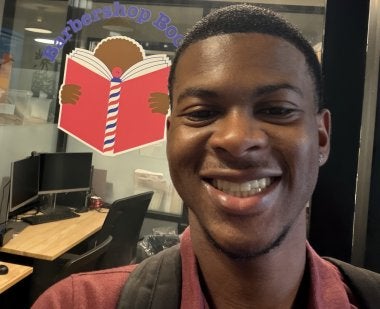 Emmanuel Dean '28 stands in front of an office with the "Barbershop Books" logo on the window, featuring a Black child reading a red book with a barbershop pole for a spine.
