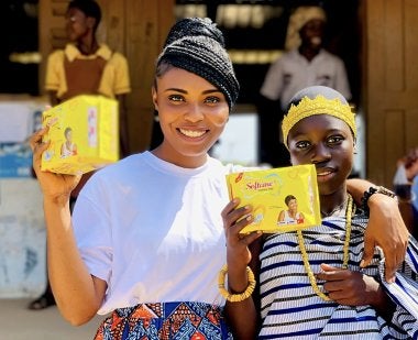 Joyce Arthur M.A. ’26, a Ph.D. student, stands with her arm around a girl in Ghana. They hold up yellow packages containing free menstrual products that Arthur distributes through her nonprofit, Araba Foundation International.