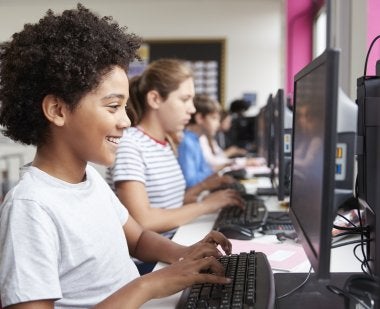 A middle school student smiles while working on a computer, in front of a line of other students at work on computers as well.