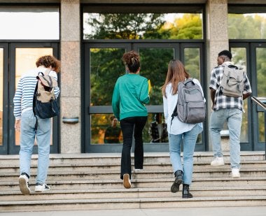 Four high school students wearing backpacks walk up steps into a school building.