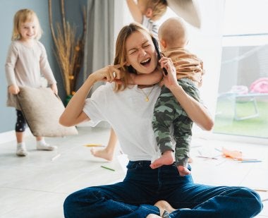 A mother puts her finger in her ear while holding a baby and a cell phone, as two children have a pillow fight in the background.