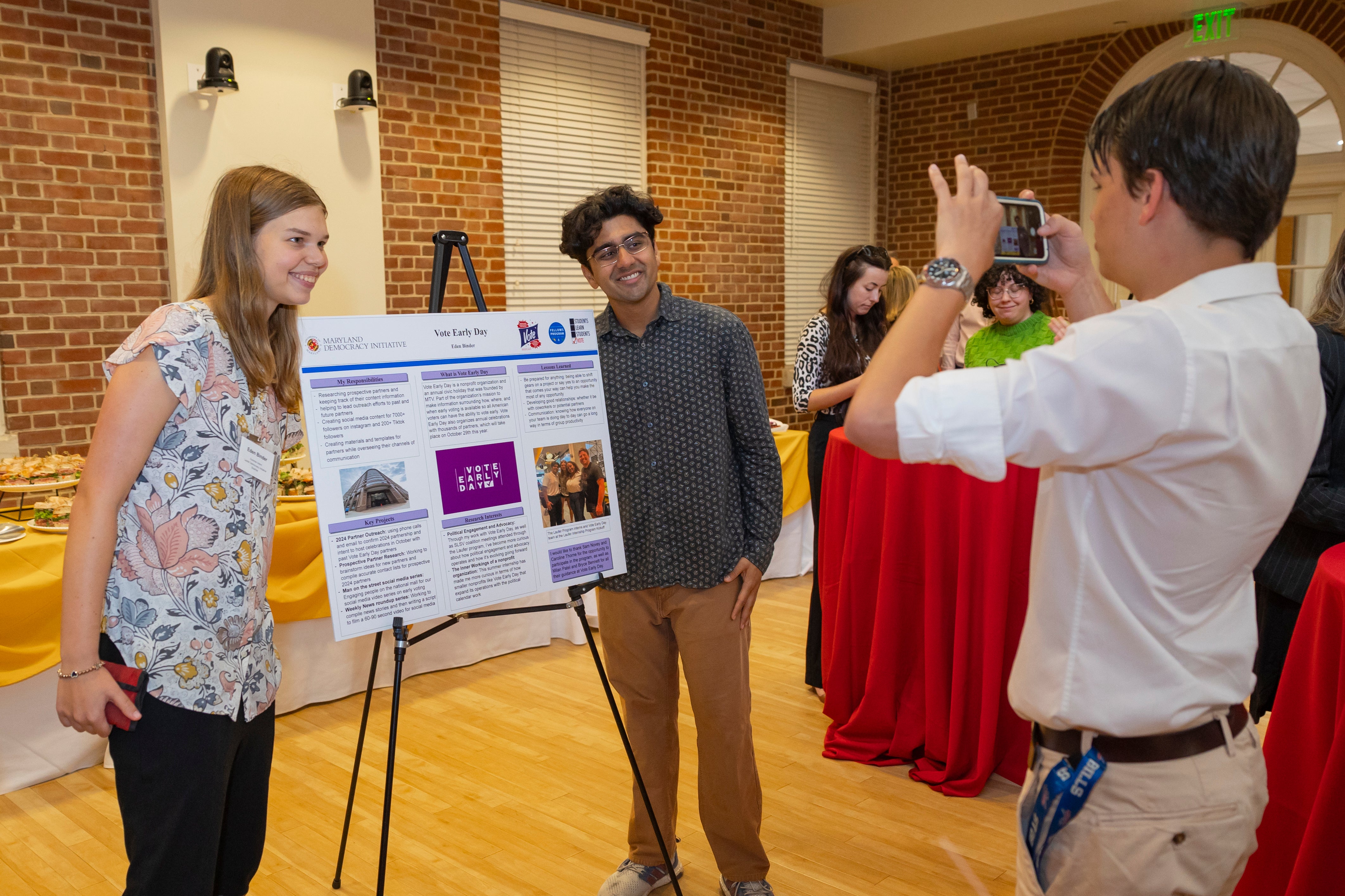 Journalism student Eden Binder (left) poses with her mentor Milan Patel, partnership manager at Vote Early Day, next to her summer poster for the Laufer Democracy Internship while journalism student Wade DeVinney (26) takes a photo. Photo by Riley N. Sims Ph.D. (23)