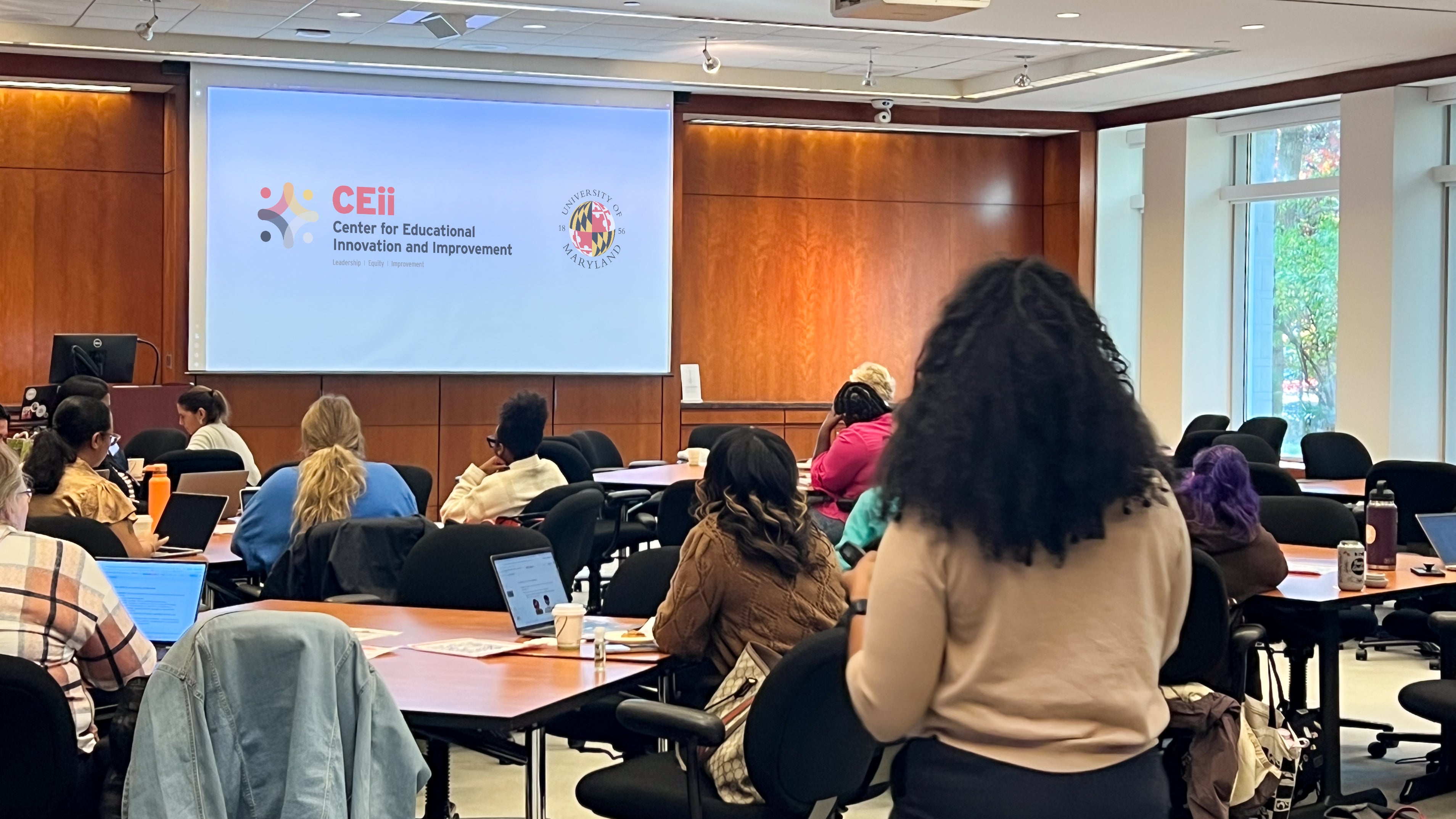 A woman stands at the back of a room at a conference speaking while a projector screen says Ceii