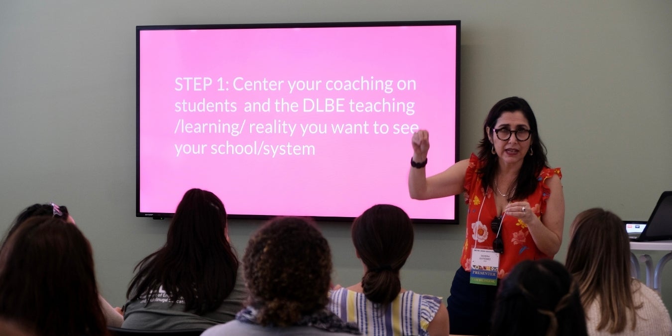 A woman speaks to a crowd in front of a screen that reads "STEP 1: center your coaching on students"