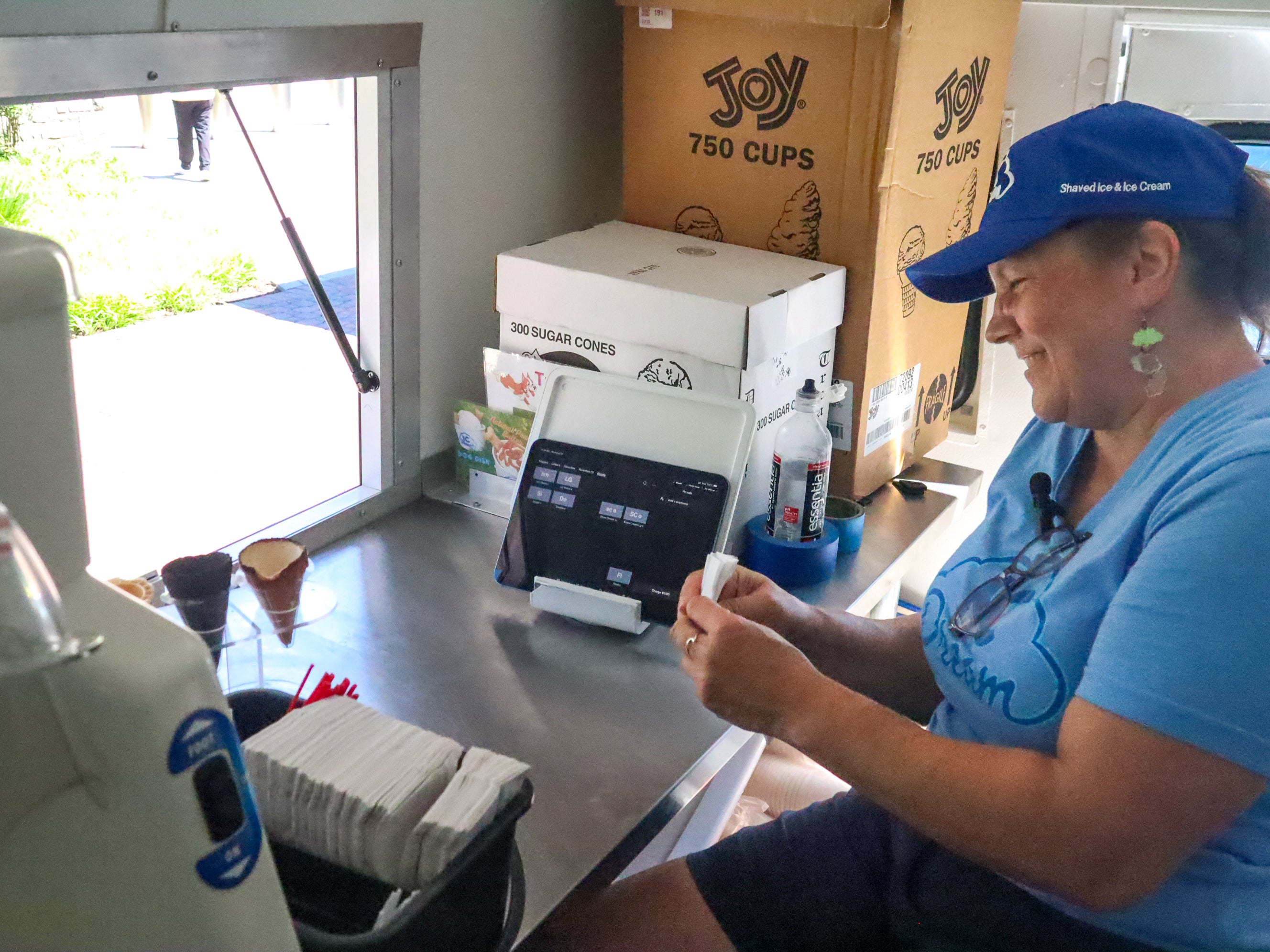 Carol Pisciotta '96 laughs as she talks to a customer from inside her ice cream truck. She is wearing a blue baseball cap and a blue shirt with her company logo on it (I C Dream).