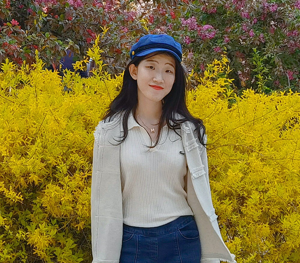 Women with long black hair, a blue hat, tan shirt standing in front of yellow flowers